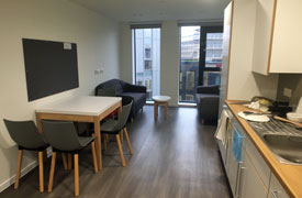 A kitchen with a sink and lots of cupboards against one wall, and a dining table with four chairs opposite. At the far end of the room there are two sofas with a small table between them.