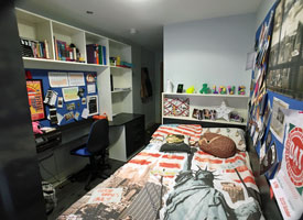 A bedroom, with bed, desk and chair, and shelving above the desk and at the end of the bed
