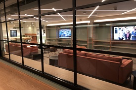 A large window in front of a room with two large televisions, two sofas, a table and several stools and armchairs.
