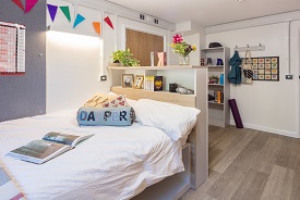 A room with a double bed in one corner, and a bookshelf and coat rack behind it.