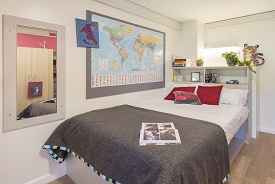 A double bed with a map of the world on the wall next to it, a mirror at the foot of the bed and a cupboard against the opposite wall.
