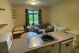 A kitchen with a sink visible, fridge and seating area in the background. A window in the far rear of the photo with some shelves in the left of the room and a microwave on the counter top.