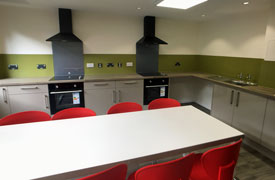 A kitchen with two ovens and hobs, two sinks, and several cupboards and drawers. There is a table in the middle of the room with seven chairs around it.