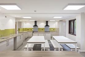 A kitchen with two ovens and hobs, three sinks, and lots of cupboards. There are two tables in the middle of the room, with two benches on either side of them.