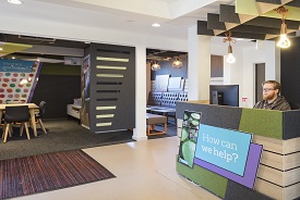 A man sat at a reception desk with the words 'how can we help?' written on it. In the background there are two rooms with chairs around tables in them.