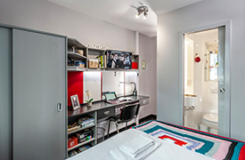 A room with a wardrobe and desk against one wall, with a chair next to the desk and a mirror, shelves and television screen over it. There is a doorway at the adjacent wall into an en suite bathroom with toilet, sink and mirror.