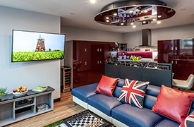 A room with a large corner sofa facing a television screen on the wall. The kitchenette area behind the sofa has several tall cupboards.