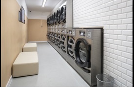 A laundry room showing a row of silver washing machines and two soft stools