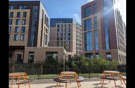 An exterior photograph of a beige brickwork building with a number of windows and doors sitting behind three orange tables
