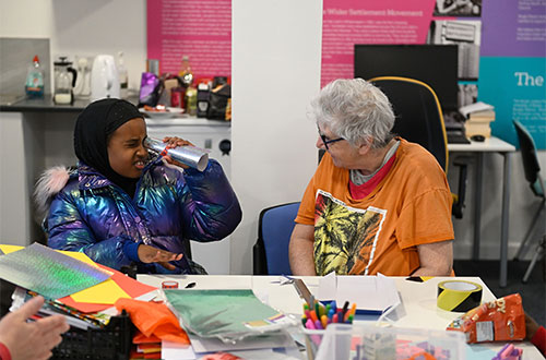 A child looking through a rolled up piece of silver card next to an adult sitting at a table with craft materials on.